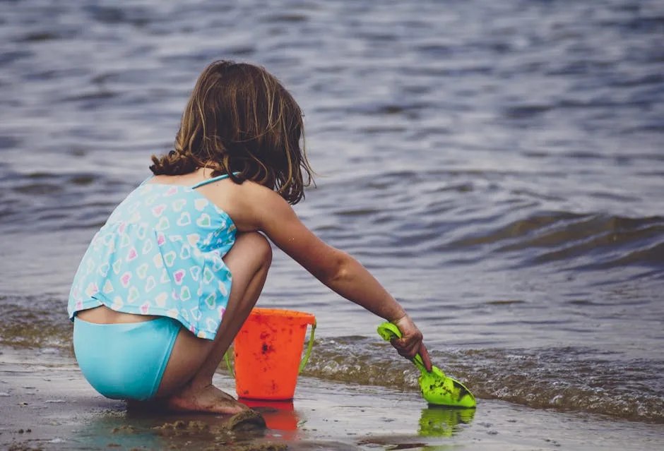 Young girl digging with green shovel beside orange bucket on beach