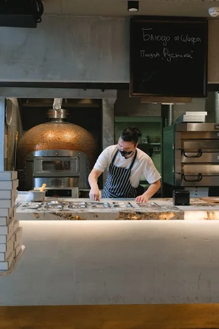 Masked chef arranging pizza orders at marble counter with wood-fired oven